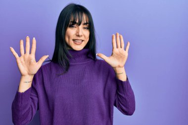 Young brunette woman with bangs wearing turtleneck sweater showing and pointing up with fingers number ten while smiling confident and happy. 