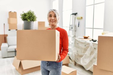 Orta yaşlı, kır saçlı, gülümseyen bir kadın yeni evinde kendine güvenen bir paket tutuyor.