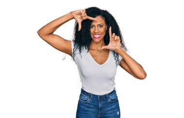 Orta yaşlı Afro-Amerikalı kadın, günlük beyaz tişört giyip gülümsüyor ve elleri ve parmakları gülen yüzlü bir çerçeve yapıyor. yaratıcılık ve fotoğrafçılık kavramı. 