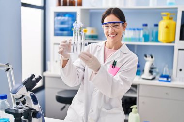 Bilim adamı üniforması giyen genç bir kadın laboratuarda elinde çiçeklerle test tüpleri tutuyor.