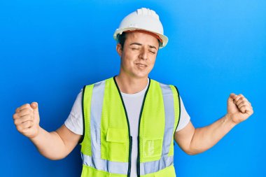 Güvenlik kaskı ve yansıtıcı ceket giyen yakışıklı genç adam mutlu ve heyecanlı bir şekilde kollarını kaldırıp, gülümseyerek ve başarı için çığlık atarak kazanan hareketi yapıyor. kutlama konsepti. 