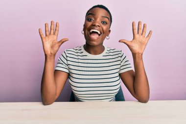 Young african american woman wearing casual clothes sitting on the table showing and pointing up with fingers number ten while smiling confident and happy. 