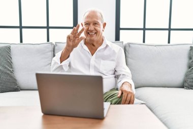 Senior man using laptop at home sitting on the sofa showing and pointing up with fingers number three while smiling confident and happy. 