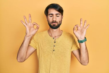 Günlük kıyafetler giyen genç İspanyol adam rahatla ve gözleri kapalı gülümse parmaklarıyla meditasyon hareketi yapıyor. yoga konsepti. 