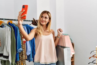 Young caucasian customer woman smiling happy holding shopping bags and make selfie by the smartphone at clothing store.
