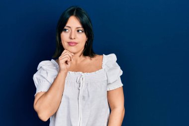 Elleri çenesinde gündelik kıyafetler giyen genç İspanyol kadın soru sormayı düşünüyor, düşünceli bir ifade. Düşünceli bir yüzle gülümsüyor. şüphe kavramı. 