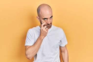 Gündelik beyaz tişört giyen genç kel adam senin jestini ve şüpheli ifadeni izliyor. 