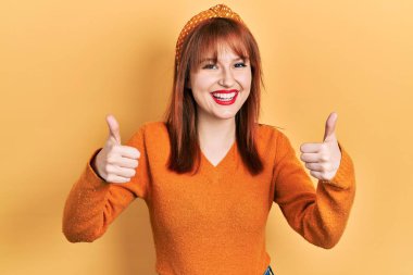 Kızıl saçlı, turuncu süveter giyen genç bir kadın. Elleriyle olumlu jest yapıyor. Gülümsüyor ve mutlu. Neşeli ifade ve kazanan jest. 