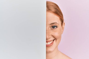 Young irish woman holding blank empty banner showing half face smiling with a happy and cool smile on face. showing teeth. 
