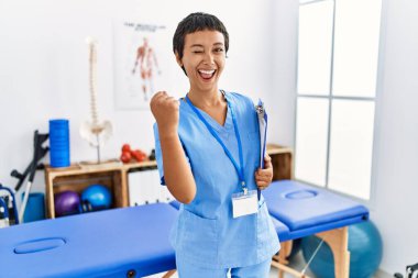 Kısa saçlı İspanyol bir kadın ağrı kesici kliniğinde çok mutlu ve heyecanlı bir şekilde çalışıyor. Kolları havada, gülümsüyor ve başarı için bağırıyor. kutlama konsepti. 