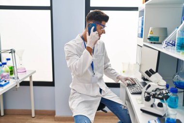 Young hispanic man wearing scientist uniform using computer and talking on the smartphone at laboratory