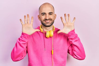 Spor kıyafetleri giyen ve kulaklık takan genç kel adam 10 numarayı gösterip işaret ederken kendinden emin ve mutlu gülümsüyor.. 