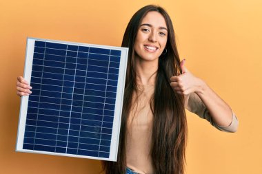 Fotovoltaik güneş paneli tutan genç İspanyol kız mutlu ve pozitif gülümsüyor, başparmağıyla mükemmel ve onay işareti yapıyor. 