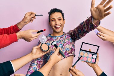 Handsome man wearing make up with make up cosmetics around looking at the camera smiling with open arms for hug. cheerful expression embracing happiness. 