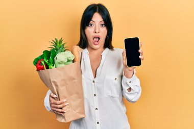 Genç İspanyol kadın elinde bir torba dolusu yiyecek, şok olmuş bir halde akıllı telefon kullanıyor, şüpheci ve alaycı görünüyor, ağzı açık kalmış. 