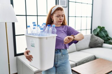 Geri dönüşüm sepetini elinde plastik şişelerle tutan genç kızıl saçlı kadın saati kontrol ediyor, rahat ve kendinden emin. 