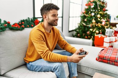 Genç Arap adam evdeki Noel ağacının yanında kanepede oturmuş video oyunu oynuyor..