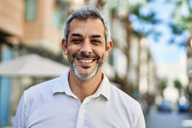 Orta yaşlı, kır saçlı, gülümseyen bir adam şehirde mutlu bir şekilde duruyor..