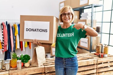 Middle age blonde woman wearing volunteer t shirt holding donations box pointing finger to one self smiling happy and proud 