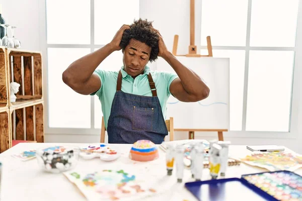 若いアフリカ系アメリカ人の男性は 痛みや片頭痛のために必死の頭痛やストレスに苦しんでアートスタジオでテーブルの上に座っています 手を頭に — ストック写真
