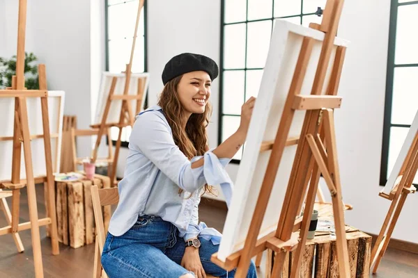若いヒスパニック系のアーティストの女性が顔に幸せとクールな笑顔でアートスタジオでキャンバスに絵を描いています 運のいい人 — ストック写真