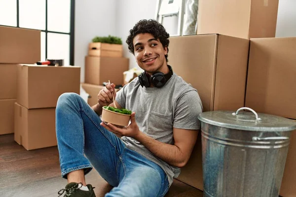 Young hispanic man eating salad sitting on the floor at new home.