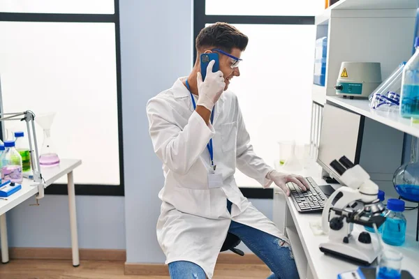 Young Hispanic Man Wearing Scientist Uniform Using Computer Talking Smartphone — стоковое фото