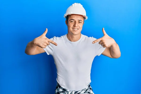 英俊的年轻人 身穿建筑工人制服 头戴安全帽 面带微笑 用手指指着自己 自豪而快乐 — 图库照片
