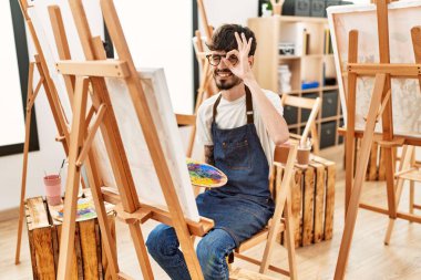 Hispanic man with beard at art studio smiling happy doing ok sign with hand on eye looking through fingers 