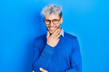 Young hispanic man with modern dyed hair wearing sweater and glasses looking confident at the camera smiling with crossed arms and hand raised on chin. thinking positive. 