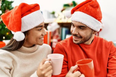 Mutlu gülümseyen ve kahve içen genç İspanyol çift evdeki Noel ağacının yanında yerde oturuyor..