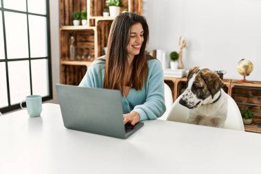 Evde köpekle aynı masada oturan dizüstü bilgisayar kullanan genç bir kadın.