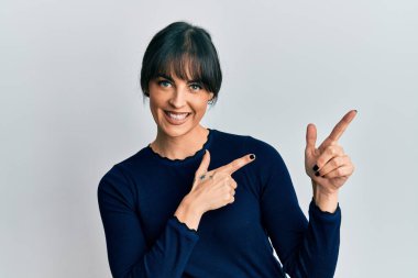 Gündelik kıyafetler giyen, gülümseyen ve kameraya bakan genç İspanyol kadın iki eliyle ve parmaklarıyla kenarı işaret ediyor.. 