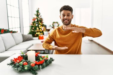 Arap genç adam noel ağacının yanında oturuyor. Elleriyle büyük ve büyük bir işaret gösteriyor. Ölçüm sembolü. Kameraya bakıp gülümsüyor. ölçüm kavramı. 