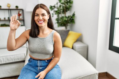 Evdeki kanepede oturan esmer kadın olumlu gülümsüyor ve el ve parmaklarıyla 