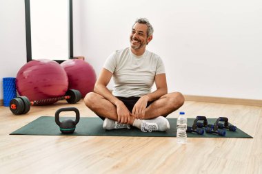 Orta yaşlı İspanyol bir adam spor salonunda antrenman minderinde oturuyor. Yüzünde gülümseme ve doğal bir ifadeyle yan yana bakıyor. Kendinden emin gülen. 