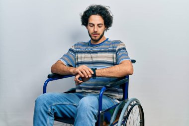 Handsome hispanic man sitting on wheelchair checking the time on wrist watch, relaxed and confident 