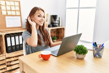 Young brunette woman working at the office with laptop smiling with hand over ear listening an hearing to rumor or gossip. deafness concept. 