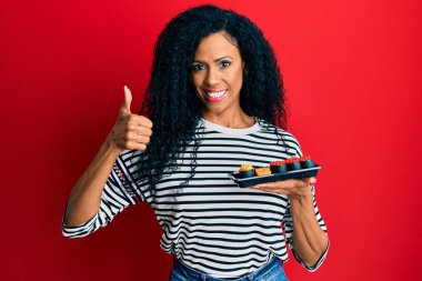 Orta yaşlı Afro-Amerikalı bir kadın bir tabak suşi gösteriyor mutlu ve pozitif gülümsüyor, başparmağıyla mükemmel ve onay işareti yapıyor. 