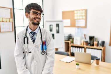 Hispanic man with beard wearing doctor uniform and stethoscope at the office looking away to side with smile on face, natural expression. laughing confident. 