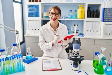 Laboratuvarda çalışan dokunmatik ped kullanan, bilim adamı üniforması giyen beyaz bir kadın.