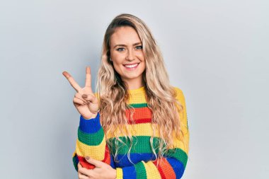 Beautiful young blonde woman wearing colored sweater smiling with happy face winking at the camera doing victory sign with fingers. number two. 