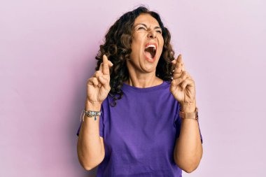 Middle age hispanic woman doing fingers crossed gesture angry and mad screaming frustrated and furious, shouting with anger looking up. 