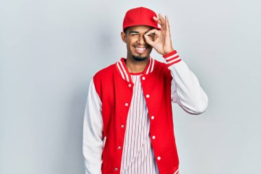 Young african american man wearing baseball uniform doing ok gesture with hand smiling, eye looking through fingers with happy face. 