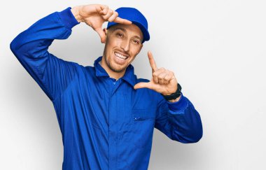 Bald man with beard wearing builder jumpsuit uniform smiling making frame with hands and fingers with happy face. creativity and photography concept. 