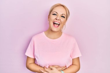 Young blonde woman wearing casual pink t shirt smiling and laughing hard out loud because funny crazy joke with hands on body. 