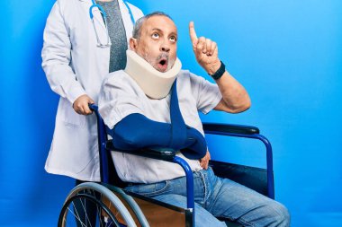 Handsome senior man with beard sitting on wheelchair with neck collar amazed and surprised looking up and pointing with fingers and raised arms. 