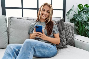 Beyaz kız akıllı telefon kullanıyor. Evdeki kanepede oturuyor..