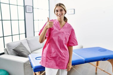 Evde fizyoterapist olarak çalışan genç sarışın kadın elleriyle mutlu bir jest yapıyor. Kameraya bakıp başarıyı gösteren ifade. 