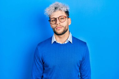 Young hispanic man with modern dyed hair wearing sweater and glasses relaxed with serious expression on face. simple and natural looking at the camera. 